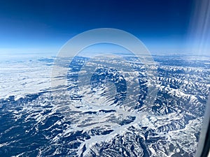 An aerial view from an airplane window of mountains, snow, clouds and blue skies