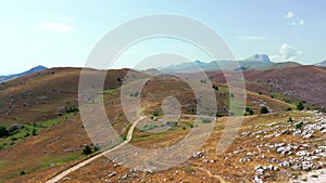 Aerial view of arid country land, some green bushes and trees, blue sky, mountain in the horizon, dirty road, white