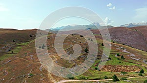 Aerial view of arid country land, some green bushes and trees, blue sky, mountain in the horizon, dirty road, white