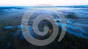 An aerial view of beautiful bog in Estonian nature during foggy sunrise with a watching tower and hiking trail, Northern Eu