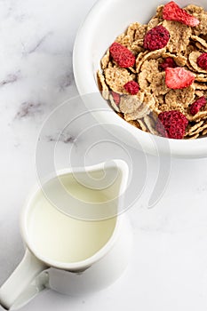 Aerial view of bowl with cereals, red berries and jug with milk, on white marble table, in vertical