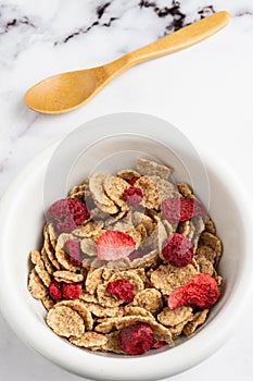 Aerial view of bowl with cereals, red berries and wooden spoon, on white marble table, in vertical