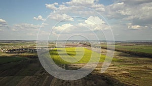 Aerial view of bright green agricultural farm field with growing rapeseed plants and distant village houses.