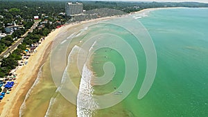 Aerial view of bustling beach with surfers catching waves, umbrellas dotting sandy shore near palm trees. Overhead drone