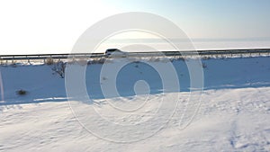 Aerial view of car riding through snow covered road near frozen lake. White SUV driving at dam route on winter day. Fly