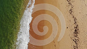 Aerial view close-up low flight over sea waves rolling on the golden sandy coast. Calm surf behind the seashell beach