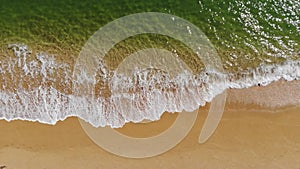 Aerial view close-up low flight over sea waves rolling on the golden sandy coast. Calm surf behind the seashell beach