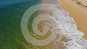 Aerial view close-up low flight over sea waves rolling on the golden sandy coast. Calm surf behind the seashell beach