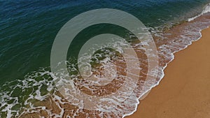 Aerial view close-up low flight over sea waves rolling on the golden sandy coast. Calm surf behind the seashell beach