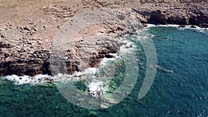 Aerial view of crystal clear waves at a rocky coast with clear blue water - camera tilt and pan