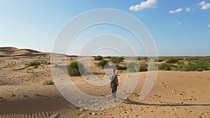 Aerial view from a drone. Woman in a long leopard dress walks along a high dune in the desert with grass in summer in