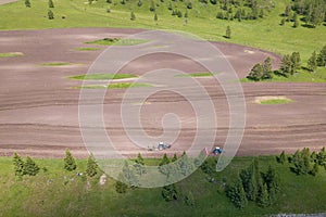 Aerial view of a farm tractor in a field during plowing of land for growing food, vegetables and fruits around the green tree with