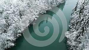 Aerial view footage with a drone moving backwards of a calm green river flowing through snow covered winter nature