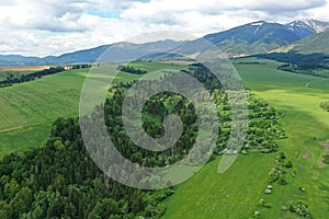 Aerial view of forested depression between two grassland fields during summer cloudy day, High Tatras mountains in background.