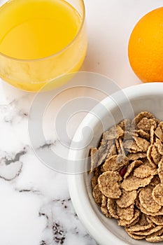 Aerial view of a glass of juice, a bowl of cereals and an orange on white marble, in vertical