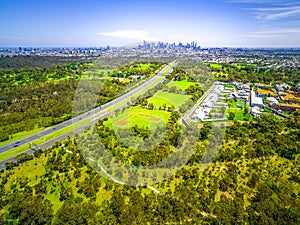 Aerial view of green parkland, Melbourne Polytechnic, and Melbourne CBD skyscrapers in the distance on summer day.