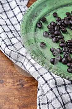 Aerial view of green plate with defocused coffee beans and defocused checkered dishcloth, on rustic wooden table
