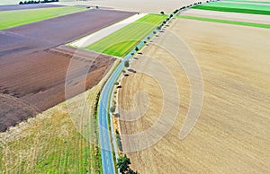 Aerial view of a grey asphalted narrow country road, which runs arcuately through a landscape with fields and meadows