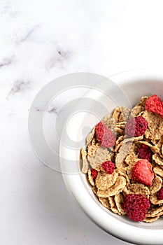 Aerial view of half a bowl with cereals and red berries on a white marble table, vertically,