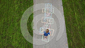 Aerial view of little boy jumping by hopscotch drawn on asphalt. Child playing hopscotch game on playground on spring day.