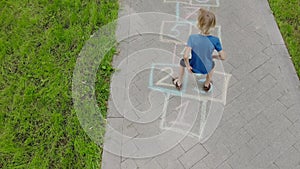 Aerial view of little boy jumping by hopscotch drawn on asphalt. Child playing hopscotch game on playground on spring day.