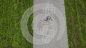 Aerial view of little boy jumping by hopscotch drawn on asphalt. Child playing hopscotch game on playground on spring day. Top vie