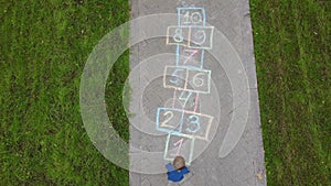 Aerial view of little boy jumping by hopscotch drawn on asphalt. Child playing hopscotch game on playground on spring day. Top vie