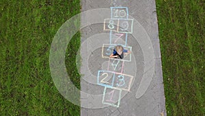 Aerial view of little boy jumping by hopscotch drawn on asphalt. Child playing hopscotch game on playground on spring day. Top vie