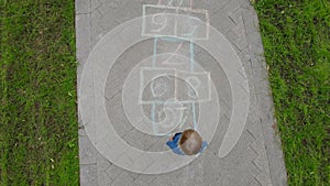 Aerial view of little boy jumping by hopscotch drawn on asphalt. Child playing hopscotch game on playground on spring day. Top vie