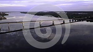 Aerial view of a long bridge above the wide river in sunset. Clip. Countryside area with a calm river near the village.