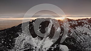 Aerial view of mountain peak during winter sunset. Snow capped mountain and orange clouds in the background.