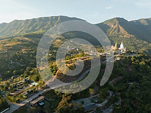 An aerial view of a mountainous landscape at sunrise, showcasing a serene temple complex with a prominent white Buddha statue