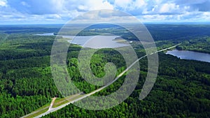 Aerial view of a narrow road becoming a bridge above the long river. Clip. Green forest and blue cloudy sky.