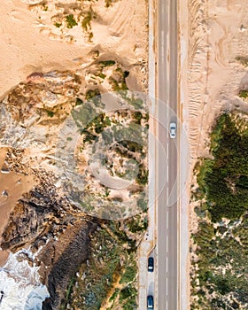 Aerial view of a narrow road with vehicles driving along the wild south coastline facing the Atlantic Ocean wit crispy waves