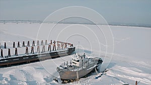 Aerial view of an old pier and a moored rusty ship on a snow covered shore. Clip. Winter landscape of an abandoned pier