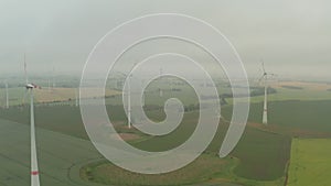 Aerial: view over foggy agricultural yellow field with multiple wind turbines generating power trough wind for a