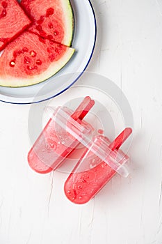 Aerial view of popsicle molds with ice cream, with plate with pieces of watermelon, vertical white background,