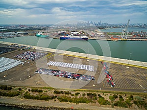 Aerial view of Port Melbourne with moored cargo vessels, imported cars parking lots, and Melbourne CBD skyline on the horizon.