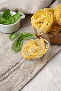 Aerial view of raw tagliatelle in wooden bowl and basil on dishcloth on white wooden table
