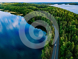 Aerial view of road with cars between green forest and blue lake water in Finland