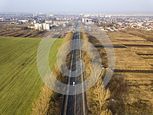 Aerial view of road with moving cars, green and plowed fields and meadow and city suburbs on sunny day. Drone photography