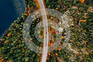 Aerial view of rural road with red car in yellow and orange autumn forest with blue lake