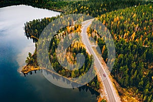 Aerial view of rural road with red car in yellow and orange autumn forest with blue lake