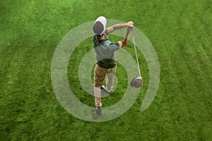 Aerial view of skilled golfer in casual attire, in cap doing completing swing in motion on green turf.