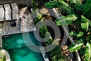 Aerial view of slim young woman in beige bikini relaxing and sunbathe near luxury swimming pool in green tropic in Ubud