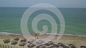 Aerial view of straw umbrellas on the beach. Ocean waves washing sandy shore. Tranquil coast line of a luxury resort.