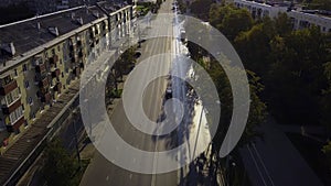 Aerial view of a sunny city street with driving cars along the alley with green trees. Clip. City in sunset time.