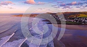 Aerial view of the town of Woolacombe and it`s beach at dawn, waves breaking on a beach, the headland of Morte Point bathed in