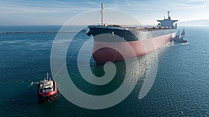 Aerial view of tug boat assisting big cargo ship. Large cargo ship enters the port escorted by tugboats