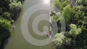 An aerial view. Two in a kayak, merging on the Amazon. The river and the banks are overgrown with trees.